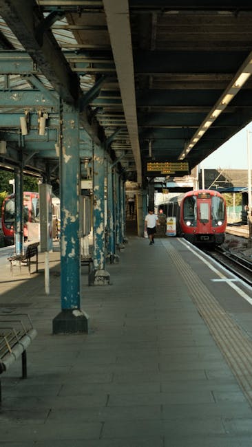 A covered train platform with weathered blue metal support beams and a wooden roof structure, where a man is seen walking along the tiled walking area beside two parked trains. One train, with a red exterior, is stationed on the right track, while the other has a white and red design. The platform is equipped with metal benches and safety line markings on the ground. The scene is lit by natural daylight, and the background shows more platforms and railway infrastructure, suggesting a busy station. The image context aligns with services related to house removals and furniture transport, as seen in representations of loading or unloading processes at a train station, relevant to relocating or moving services provided by Man with Van Barnet.
