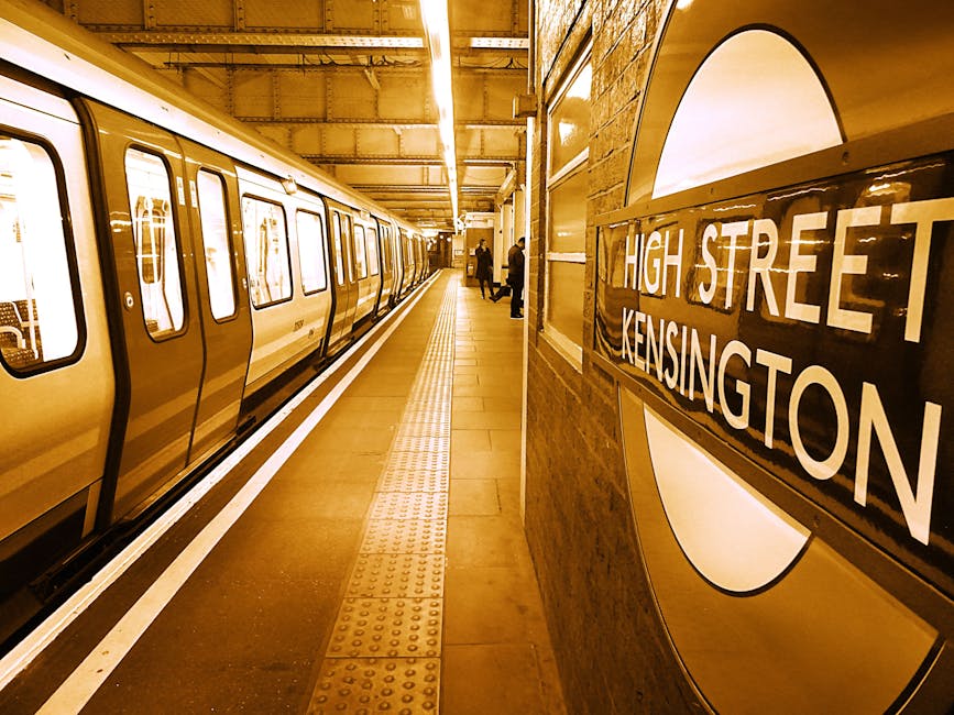 Photograph of a London Underground station platform at High Street Kensington, showing a packed train on the left with closed doors and visible window reflections. The platform is illuminated with warm lighting, and the tiled floor features tactile paving strips near the edge for safety. Several passengers stand at a distance, some facing the train and others walking along the platform. On the right side, there is a brick wall with a roundel sign displaying 'HIGH STREET KENSINGTON' in white letters on a black background. The station environment appears clean and well-maintained, with equipment and signage visible, providing a typical setting for tube travel in London. This image reflects the busy atmosphere experienced during daily home relocations or moving logistics involving tube transport, as managed by local services such as Man with Van Barnet.