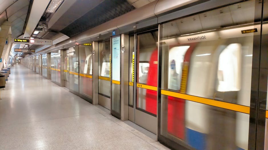 A covered train platform with weathered blue metal support beams and a wooden roof structure, where a man is seen walking along the tiled walking area beside two parked trains. One train, with a red exterior, is stationed on the right track, while the other has a white and red design. The platform is equipped with metal benches and safety line markings on the ground. The scene is lit by natural daylight, and the background shows more platforms and railway infrastructure, suggesting a busy station. The image context aligns with services related to house removals and furniture transport, as seen in representations of loading or unloading processes at a train station, relevant to relocating or moving services provided by Man with Van Barnet.