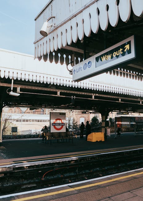 A clear daytime view of New Barnet station with a bright blue sky and scattered white clouds overhead. In the foreground, there is a spacious paved area where several people are standing or walking, s